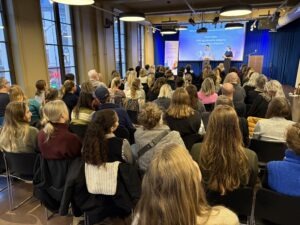 Mange personer som sitter i et rom mot en scene. På scenen står det to konferansierer.